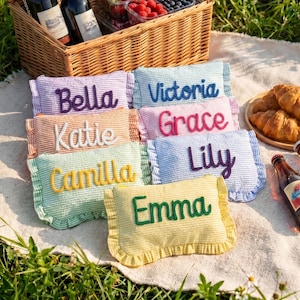Könnte beinhalten: Sieben rechteckige Stoffbeutel mit Rüschen in verschiedenen Pastellfarben, jeweils mit einem individuellen Namen bestickt: Bella, Victoria, Katie, Grace, Camilla, Lily und Emma. Die Artikel liegen auf einem weißen Tuch neben einem Picknickkorb.