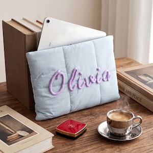 May include: A light blue quilted laptop sleeve featuring the name Olivia embroidered in purple script lettering on the front. The rectangular sleeve is constructed with vertical stitching and is designed to hold a tablet or laptop securely.