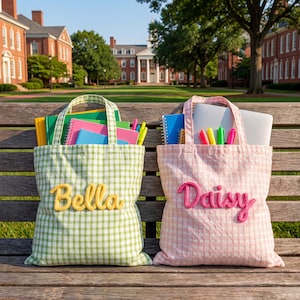 May include: Two fabric tote bags with a gingham pattern, one in light green and one in light pink. The green bag features the name Bella in yellow yarn, and the pink bag features the name Daisy in pink yarn. Both bags are filled with notebooks and pens.