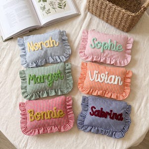 May include: Six rectangular zipped pouches made of gingham fabric with ruffled edges. Each pouch features a different colour and a personalised name embroidered in a raised, cursive font: Norah, Sophie, Margot, Vivian, Bonnie, and Sabrina.