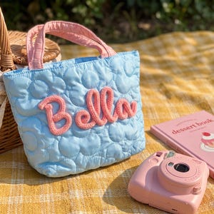 May include: A light blue quilted tote bag featuring a repeating flower pattern and the name Bella embroidered in pink yarn across the front. The bag has two pink fabric handles and a soft, padded texture throughout its exterior surface.