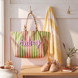 May include: A tote bag featuring vertical pink and green stripes with the name Audrey in raised lavender script lettering. The bag has two matching striped shoulder straps and is hanging on a hook next to a yellow and white striped towel.