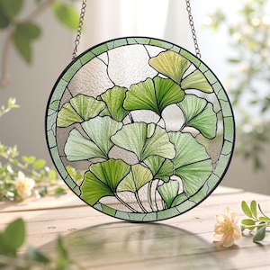 May include: Round stained glass window with a black border and chain for hanging. The design features green ginkgo leaves in varying shades, creating a nature-inspired decorative piece. The glass has a textured background.