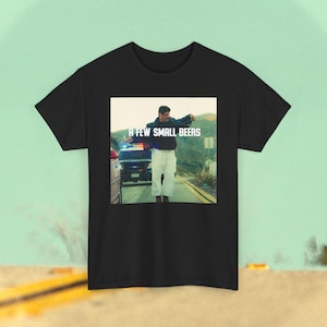 May include: A black short-sleeved t-shirt featuring a centered graphic print of a person standing on a road with a police car in the background. The text A FEW SMALL BEERS is printed in white capital letters across the center of the graphic image.