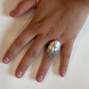 May include: Silver-toned ring with a large, textured oval design. The ring's surface has a hammered appearance, reflecting light. The ring is worn on a finger, showcasing its size and detail. A piece of jewelry.