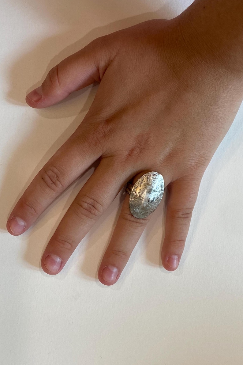 May include: Silver-toned ring with a large, textured oval design. The ring's surface has a hammered appearance, reflecting light. The ring is worn on a finger, showcasing its size and detail. A piece of jewelry.