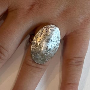 May include: A silver-toned ring with an oval, hammered design. The ring is worn on a finger, showcasing the textured surface and metallic sheen. The ring's design is simple yet eye-catching, suitable for everyday wear.