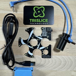 Può includere: Un set di componenti elettronici per un progetto fai-da-te. Il set include una scheda a circuito stampato nera con segni bianchi e neri, una scatola nera con un connettore USB, una scatola nera con la parte superiore blu, due cavi blu con connettori neri e una carta bianca e nera con il testo "TRISLICE TECHNOLOGY".
