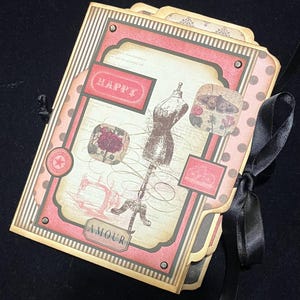 May include: A decorative scrapbook album with a black satin ribbon closure. The cover features a pink and cream color scheme with vintage-style illustrations, including a dress form, flowers, and the word "HAPPY".