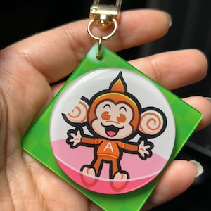 Könnte beinhalten: Ein Schlüsselanhänger mit einem Cartoon-Affen mit herzförmigen Augen, der ein orangefarbenes Outfit mit dem Buchstaben "A" trägt. Der Affe befindet sich auf einem weißen und rosa kreisförmigen Hintergrund, der auf einem grünen Quadrat mit einem goldenen Verschluss angebracht ist.