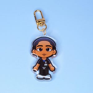 May include: A clear acrylic keychain featuring a cartoon illustration of a healthcare worker in scrubs. The keychain has a gold-colored clasp and a blue background. The healthcare worker has brown skin, dark hair, and is wearing a black uniform.