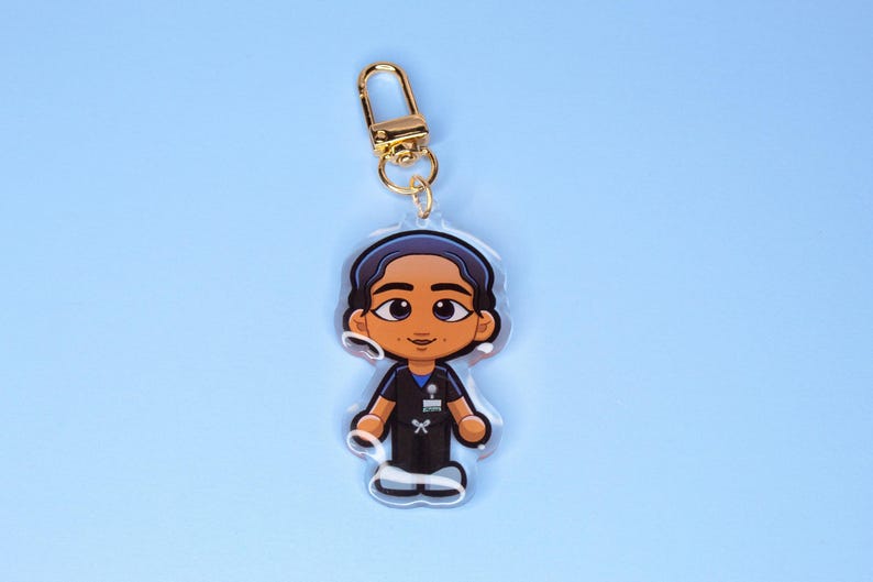 May include: A clear acrylic keychain featuring a cartoon illustration of a healthcare worker in scrubs. The keychain has a gold-colored clasp and a blue background. The healthcare worker has brown skin, dark hair, and is wearing a black uniform.