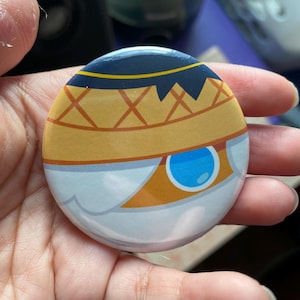 May include: A round button with a white, blue, and orange design. The design features a stylized eye with a blue iris and a yellow sclera. The eye is surrounded by a white and orange pattern.