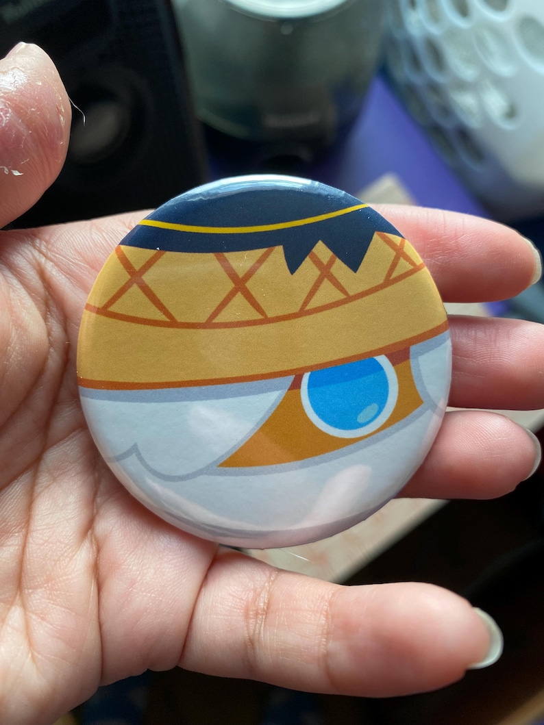 May include: A round button with a white, blue, and orange design. The design features a stylized eye with a blue iris and a yellow sclera. The eye is surrounded by a white and orange pattern.