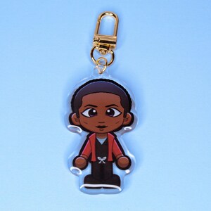 May include: A clear acrylic keychain featuring a cartoon character with short dark hair, wearing a red jacket over a black shirt and black pants. The character has large eyes and is attached to a gold-colored metal lobster clasp.