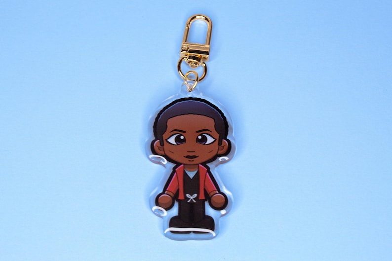 May include: A clear acrylic keychain featuring a cartoon character with short dark hair, wearing a red jacket over a black shirt and black pants. The character has large eyes and is attached to a gold-colored metal lobster clasp.