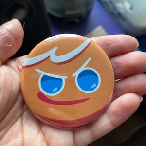 May include: A round button with a cartoon gingerbread cookie face. The cookie has a mischievous expression with blue eyes and a red smile.