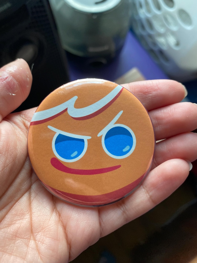 May include: A round button with a cartoon gingerbread cookie face. The cookie has a mischievous expression with blue eyes and a red smile.