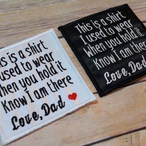 Peut inclure: Deux écussons brodés, un blanc et un noir, avec le même texte en écriture blanche : "C'est une chemise que je portais quand tu la tiens, sache que je suis là, Love, Dad." Chacun a un petit cœur rouge.