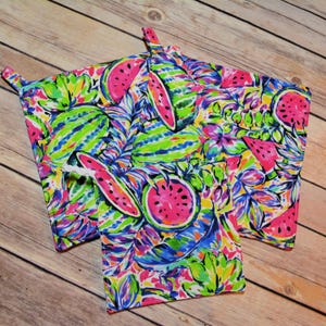 May include: Set of three colorful kitchen pot holders with a watermelon and tropical leaf pattern. The pot holders are made of fabric and have a white trim.