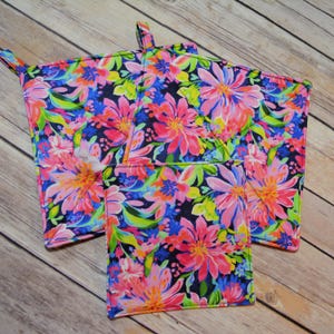 May include: Set of three colorful floral print pot holders with a dark blue background. The pot holders have a loop for hanging.