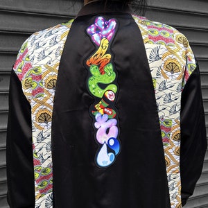 Könnte beinhalten: Schwarze Satin-Bomberjacke mit farbenfrohem Grafikdesign auf der Rückseite. Das Design zeigt eine vertikale Reihe von bunten Buchstaben und Symbolen. Die Jacke hat einen weißen und gelben gemusterten Besatz an den Ärmeln und Seiten.