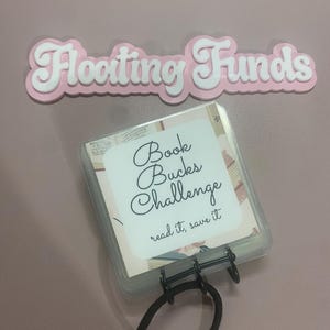 May include: A pink and white sign that reads "Floating Funds" is above a clear plastic case with a black elastic band. The case contains cards labeled "Book Bucks Challenge" with the text "read it, save it."