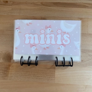 May include: A pink binder with the word "minis" in a retro font. The cover features a pattern of white geese with pink bows and flowers. The binder has black metal rings.