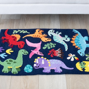 May include: A rectangular, navy blue rug featuring colorful, cartoon-style dinosaurs. The rug has a green, red, orange, blue, and purple dinosaur design, along with other decorative elements. The rug is placed on a light wood floor.