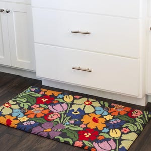May include: A rectangular floral rug with a dark background and a vibrant array of flowers in red, orange, yellow, purple, and blue. The rug is placed in front of white kitchen cabinets with silver handles.