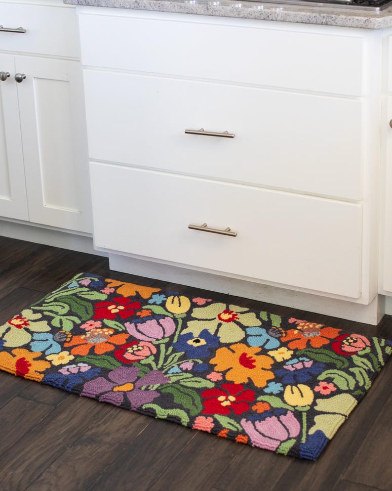 May include: A rectangular floral rug with a dark background and a vibrant array of flowers in red, orange, yellow, purple, and blue. The rug is placed in front of white kitchen cabinets with silver handles.
