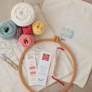 May include: A punch needle embroidery kit with a white canvas, a wooden hoop, a punch needle, a stitch gauge, and five balls of yarn in various colors: light blue, brown, coral, white, and yellow.