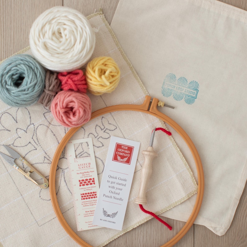 May include: A punch needle embroidery kit with a white canvas, a wooden hoop, a punch needle, a stitch gauge, and five balls of yarn in various colors: light blue, brown, coral, white, and yellow.
