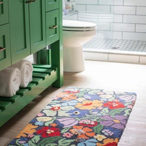 May include: A rectangular floral bath mat with a dark blue background and colorful flowers in red, orange, purple, and yellow. The mat is placed on a light wood floor in a bathroom setting.