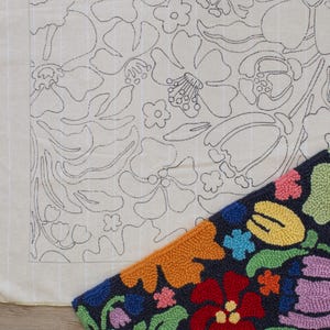 May include: A close-up of two textile pieces. One is a cream-colored fabric with a floral outline design in black. The other is a completed rug with a dark blue background and colorful, textured floral patterns in red, orange, yellow, pink, and purple.