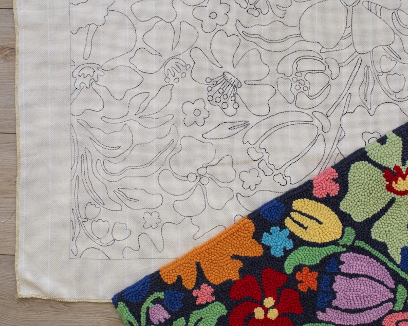 May include: A close-up of two textile pieces. One is a cream-colored fabric with a floral outline design in black. The other is a completed rug with a dark blue background and colorful, textured floral patterns in red, orange, yellow, pink, and purple.