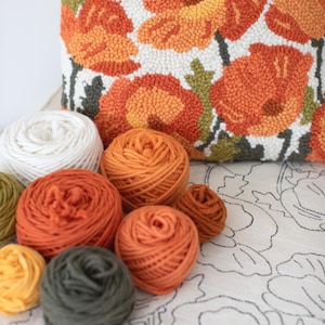 May include: A close-up of a variety of yarn balls in different colours, including white, orange, and green. The yarn balls are arranged on a white surface with a floral pattern.