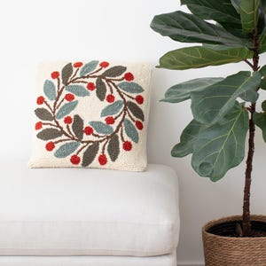 May include: Square cream-colored decorative pillow with a wreath design. The wreath features red berries, gray-brown stems, and blue-green leaves. The pillow is placed on a white surface, with a large green plant in the background.