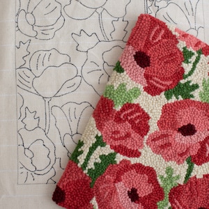 May include: A close-up of a hand-stitched rug with a floral design. The rug features a pattern of red poppies with green leaves on a cream background. The rug is partially covering a piece of fabric with a black outline of the same poppy design.