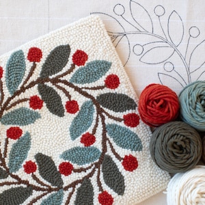 May include: A finished, off-white, tufted rug featuring a wreath design with red berries, grey and blue-green leaves, and brown stems. Several balls of yarn in red, grey, blue-green, and white are next to the rug. A canvas with a leaf outline is in the background.