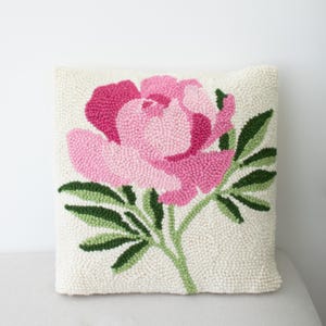 May include: Square ivory pillow with a large, embroidered peony. The flower is a gradient of pink hues, with green leaves and stem. The pillow is a decorative home accent.