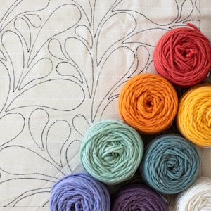 Op de afbeelding: Een verzameling van acht garenbollen in verschillende kleuren, waaronder oranje, geel, groen, blauw, paars en wit. De garenbollen zijn in een halve cirkel gerangschikt op een witte stoffen achtergrond met een bloemenpatroon.