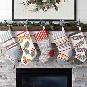 May include: Five Christmas stockings with various patterns hang above a fireplace. The stockings feature designs like stripes, snowflakes, holly, and geometric shapes, in colors of red, green, white, and blue. A garland and artwork complete the festive scene.