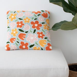Op de afbeelding: Een vierkant kussen met een bloemenpatroon. Het kussen is gemaakt van een zachte, pluche stof en heeft een verscheidenheid aan kleurrijke bloemen in tinten oranje, geel, roze en groen. De achtergrond van het kussen is lichtblauw.