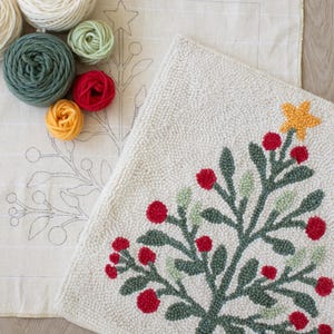 May include: A cream-coloured, hand-stitched Christmas tree rug with a star and red berry accents. Beside it, a canvas with a tree outline and various yarn skeins in cream, green, red, and yellow. A craft project.