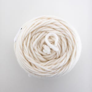 May include: A top-down view of a tightly wound ball of off-white yarn. The yarn is thick and appears soft, with a loop of yarn visible at the center. The background is plain white, highlighting the texture and color of the yarn.