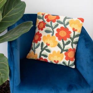 May include: A white throw pillow with a floral pattern featuring orange, yellow, and green flowers. The pillow is sitting on a blue velvet chair.
