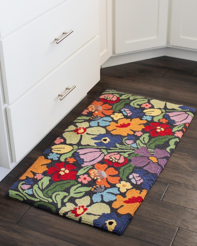 May include: A rectangular floral rug with a dark background and a vibrant array of flowers in red, orange, yellow, blue, and purple. The rug is placed on a dark wood floor near white cabinets.