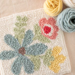 May include: A close-up of a floral punch needle project with a blue flower, pink flower, and yellow flower. The project is surrounded by balls of yarn in various colors.