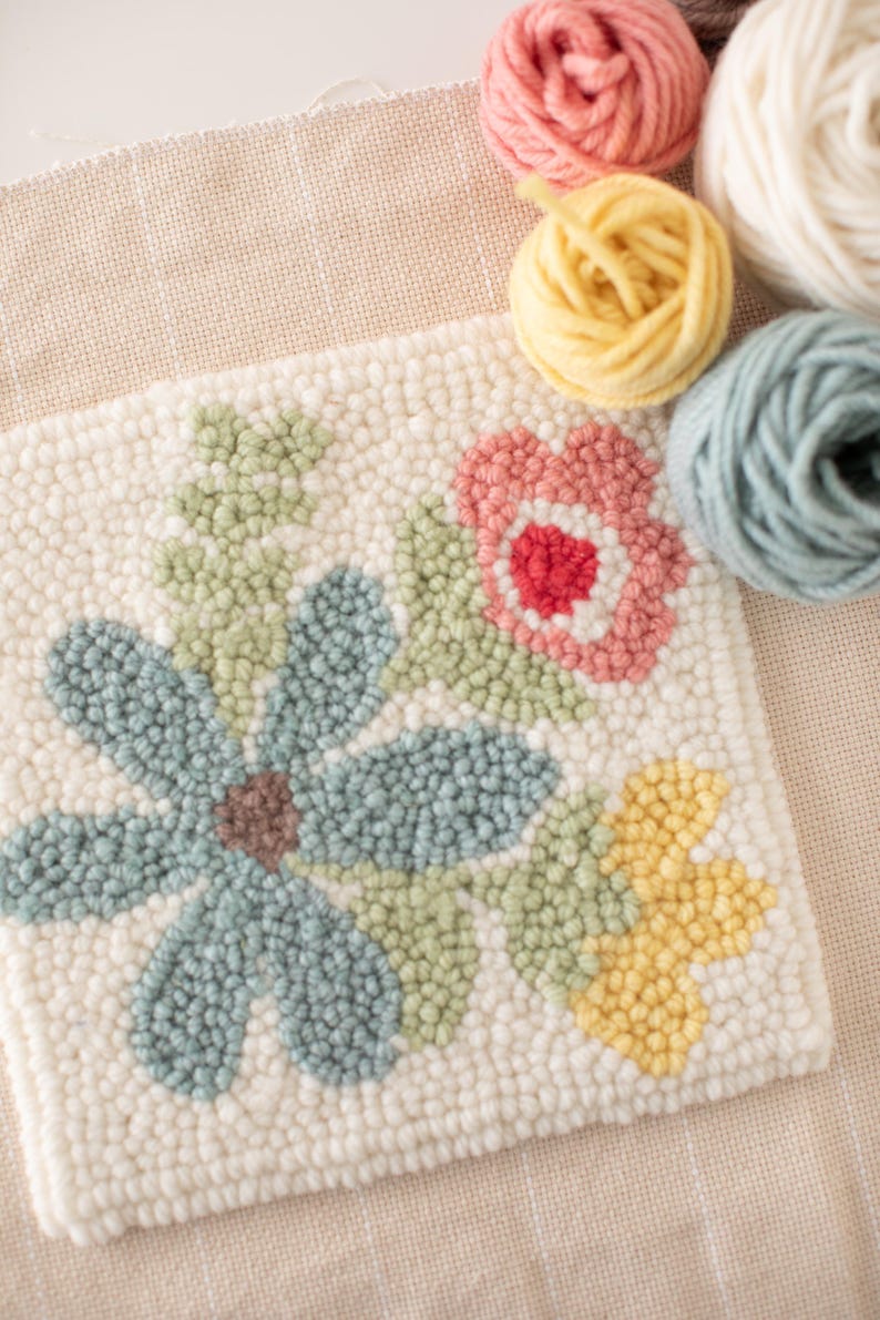 May include: A close-up of a floral punch needle project with a blue flower, pink flower, and yellow flower. The project is surrounded by balls of yarn in various colors.